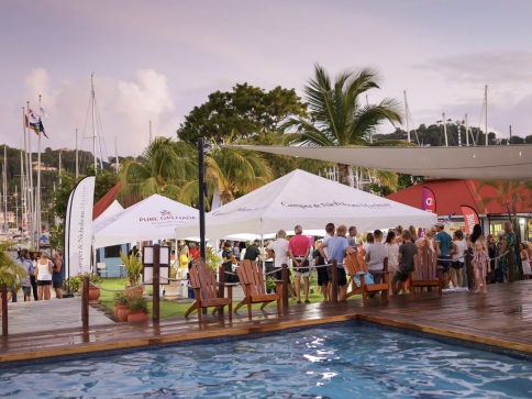 A group of people gather under white marquees beside a swimming pool and wooden decking at a marina, with sailing boats, palm trees, and flags visible in the background at sunset.
