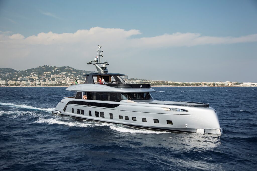 A sleek, modern yacht with multiple decks cruises on deep blue water near a coastal city with many buildings and green hills in the background under a partly cloudy sky.