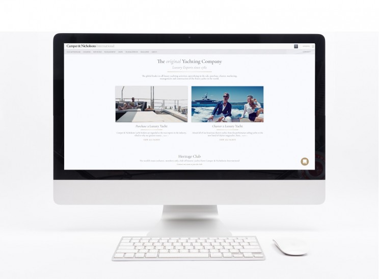 A computer monitor displaying a yachting company’s website with images of people on luxury yachts, placed on a white desk with a keyboard and mouse in front.