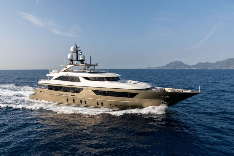 A large, modern luxury yacht with a sleek design sails on deep blue water, leaving a white wake behind. The sky is clear, and distant hills are visible on the horizon.