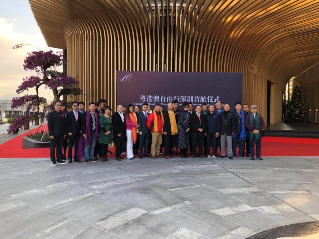 A group of people pose for a photo in front of a large sign at an outdoor event with modern wooden architecture and a red carpet. Some are wearing scarves and coats. There is a decorated tree on the left.