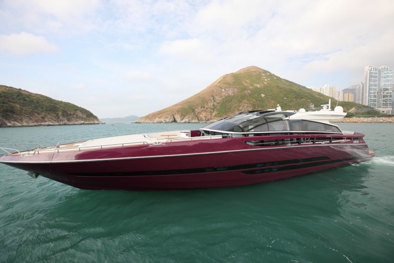 A sleek, burgundy luxury yacht cruises on turquoise water with green hills and modern buildings in the background under a partly cloudy sky.