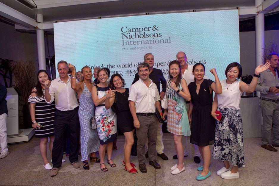 A group of people pose and smile in front of a Camper & Nicholsons International sign at an indoor event, with several individuals waving at the camera.