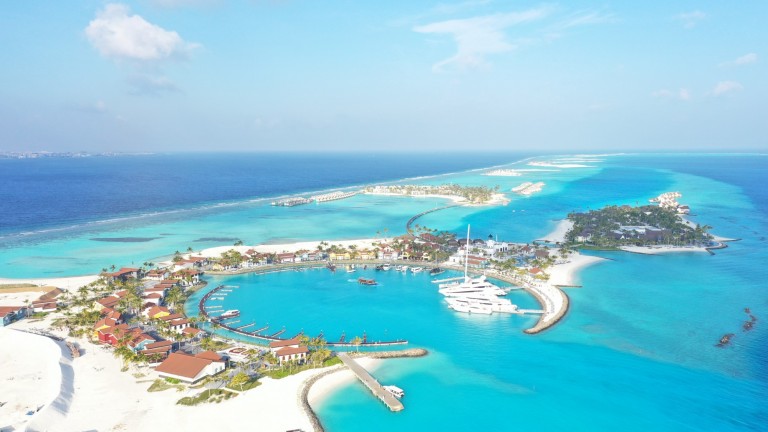 Aerial view of a tropical island resort with turquoise waters, white sandy beaches, boats moored at a marina, and overwater villas surrounded by vibrant coral reefs under a clear blue sky.