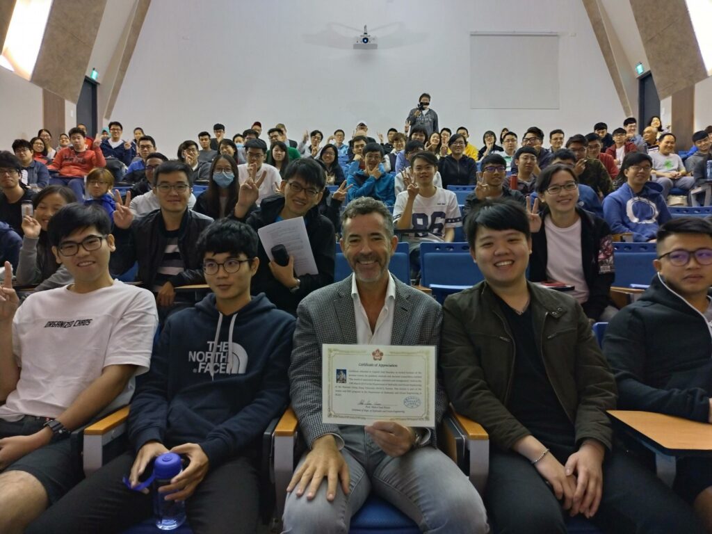 A group of students sits in an auditorium, smiling at the camera. A man in a grey jacket sits front and centre, holding a certificate. The setting appears to be a classroom or lecture theatre.
