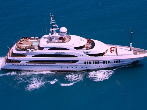 A large, luxurious white yacht cruises through deep blue water, leaving a trail of white waves behind. The yacht has multiple decks with outdoor seating, a hot tub, and a helipad.