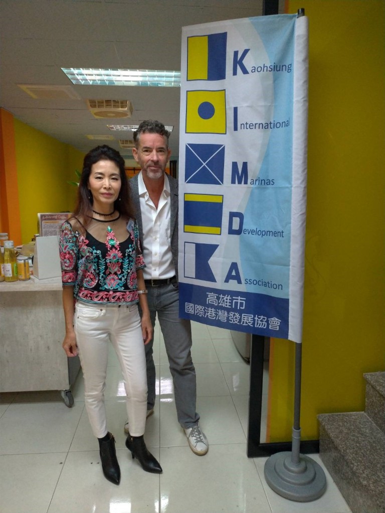 A woman and a man stand indoors next to a banner for the Kaohsiung International Marinas Development Association, featuring blue and yellow nautical flag designs. The room has yellow walls and various items on a table behind them.
