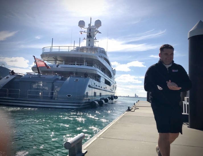 A man in a black outfit walks on a sunlit quay next to a large, modern yacht moored in clear blue water, with city buildings visible in the distant background.