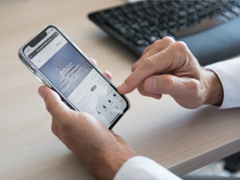 A person holds a mobile phone in one hand while using the other hand to navigate a yacht booking app; a keyboard is visible on the desk in the background.