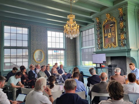 A group of people sits in a stylish, ornate room with large windows, a chandelier, and a decorative wall, attentively watching a presentation on a screen at the front.