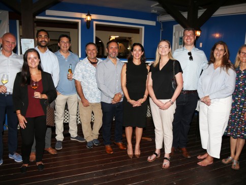 A group of eleven adults, dressed in smart casual attire, pose and smile together indoors on a wooden floor with blue walls in the background. Some are holding drinks, and the setting appears to be a social or professional gathering.