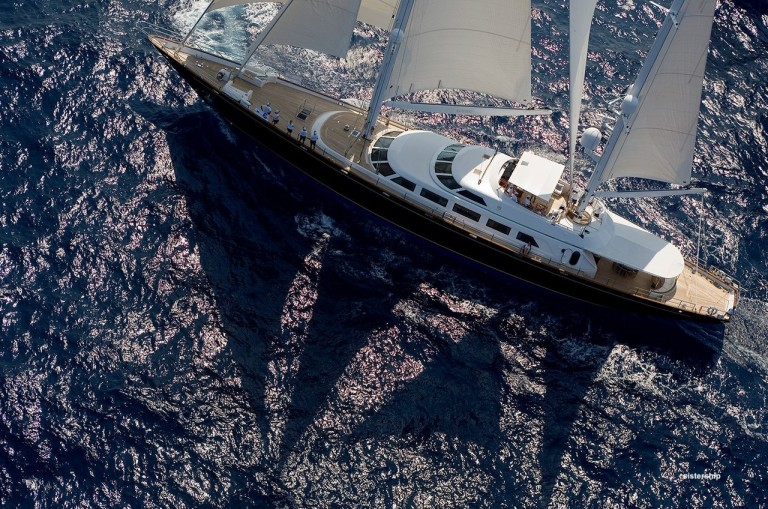 Aerial view of a large sailing boat with white sails gliding over deep blue ocean water, casting distinct shadows on the waves.