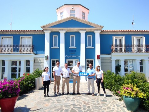 Six people stand smiling in front of a blue and white building with a sign reading Capitainerie. One person holds a blue 2023 sign. Colourful flowers and green bushes line the walkway to the entrance.