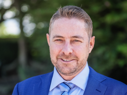 A man with short light brown hair and a trimmed beard, wearing a blue suit, light blue shirt, and striped tie, smiles at the camera outdoors with greenery in the background.