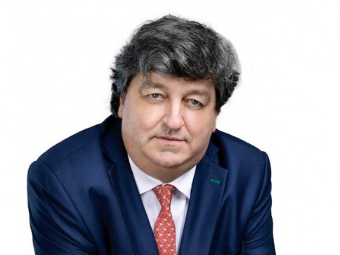 A middle-aged man with grey, wavy hair wearing a blue suit, white shirt, and red patterned tie poses against a plain white background.