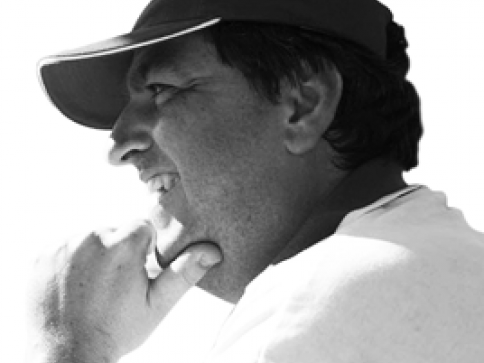 A man wearing a cap looks to the left and smiles, resting his chin on his hand. The photo is in black and white and shows a close-up side profile of his face.