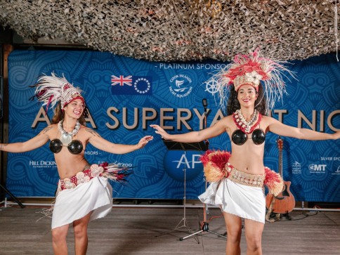 Two women in traditional Polynesian attire perform a dance on stage. They wear decorative headdresses, coconut-shell tops, and white skirts. A blue event banner and musical instruments are visible in the background.