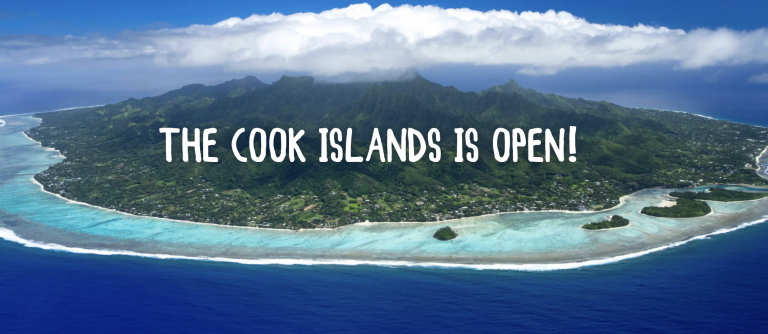 Aerial view of a lush green island surrounded by turquoise waters and a coral reef, with white clouds over the mountains. Bold text reads, THE COOK ISLANDS ARE OPEN! across the centre.