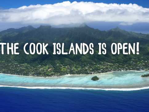 Aerial view of the lush, green Cook Islands surrounded by turquoise waters and a reef, with “THE COOK ISLANDS ARE OPEN!” written in bold white letters across the image.