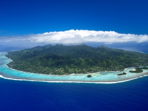 Aerial view of a lush green island surrounded by turquoise water and coral reefs, with a line of clouds covering the mountain peaks under a clear blue sky.