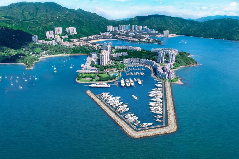 Aerial view of a marina filled with yachts and boats, surrounded by breakwaters, with residential buildings, green hills, and mountains in the background under a blue sky.