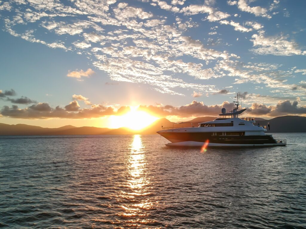 A luxury yacht is anchored on calm water during sunset, with the sun low on the horizon, casting a golden reflection on the water and clouds scattered across the sky.