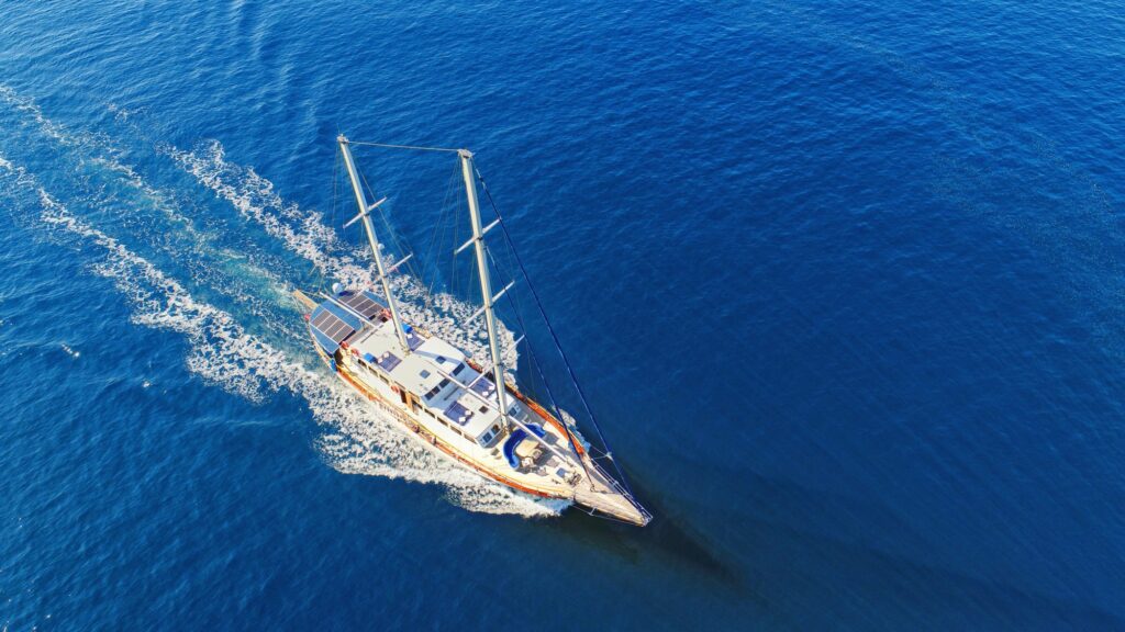 Aerial view of a white sailing boat with two masts moving through calm, deep blue ocean water, leaving a trail of ripples behind it.
