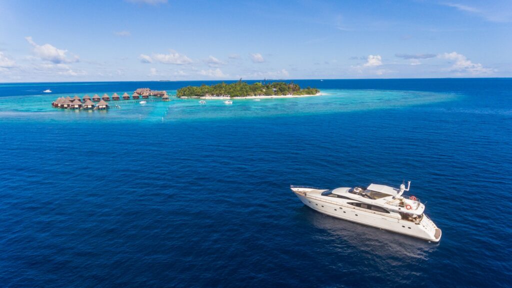 A white yacht floats on deep blue ocean water near a tropical island with overwater bungalows, palm trees, and clear skies overhead.