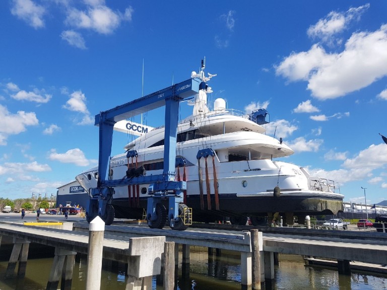 A large white yacht is lifted out of the water by a blue travel lift at a marina under a bright blue sky with scattered clouds. The scene includes pontoons and the marina facility in the background.