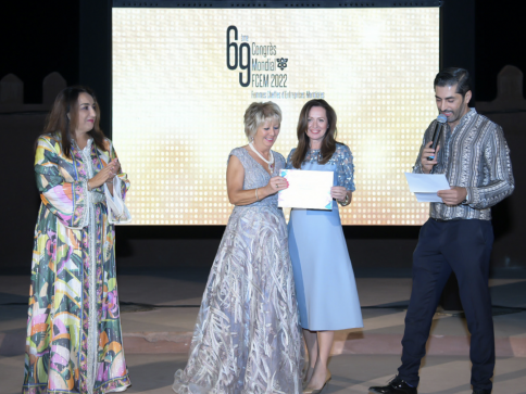 Four people stand on stage at an event, with two women in the centre holding certificates and smiling. A man on the right speaks into a microphone, while another woman on the left watches. A large event screen is behind them.