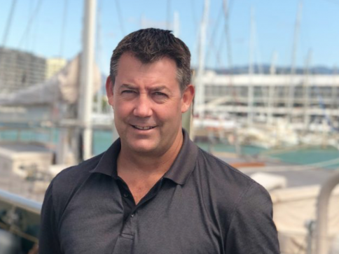 A man in a dark polo shirt stands outdoors in front of a harbour with boats and masts, under a clear sky.
