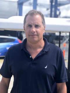 A man with short brown hair, wearing a navy blue polo shirt, stands outdoors near a blue car and some industrial equipment, under partly cloudy skies.