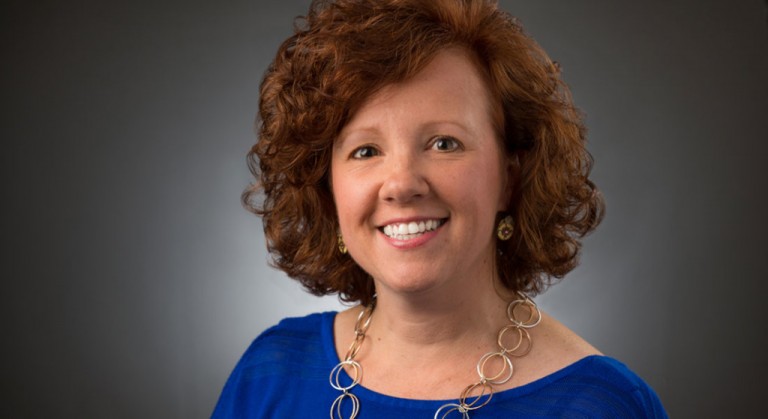 A woman with curly red hair smiles at the camera. She is wearing a royal blue top, gold hoop earrings, and a gold necklace with large circular links. The background is a plain grey gradient.