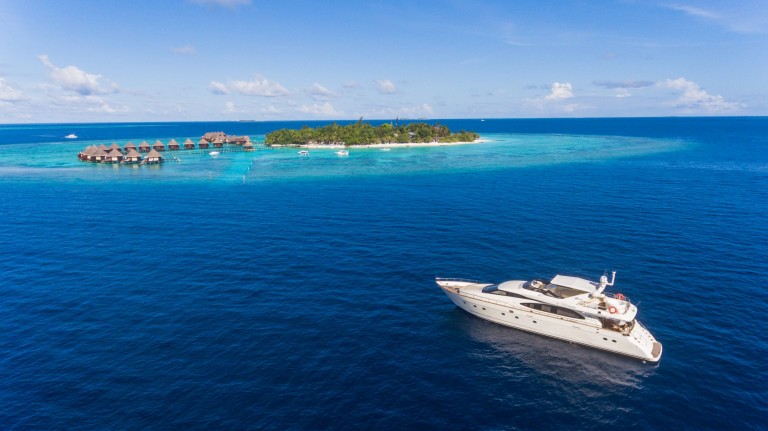 A white yacht floats on deep blue water near a lush tropical island with overwater bungalows, palm trees, and clear turquoise shallows under a bright, partly cloudy sky.