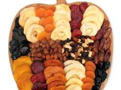 A wooden apple-shaped tray filled with assorted dried fruits—apricots, apples, bananas, plums, and more—along with sections of nuts, arranged in neat rows on a white background.