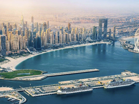 Aerial view of Dubai Marina with tall skyscrapers lining the waterfront, a curved sandy beach, and cruise ships moored at the marina pier under a hazy sky.