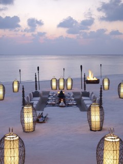 A romantic seaside dining setup on the sand at sunset, with lanterns, low tables, floor cushions, and a firepit, overlooking a calm sea and a cloudy sky.
