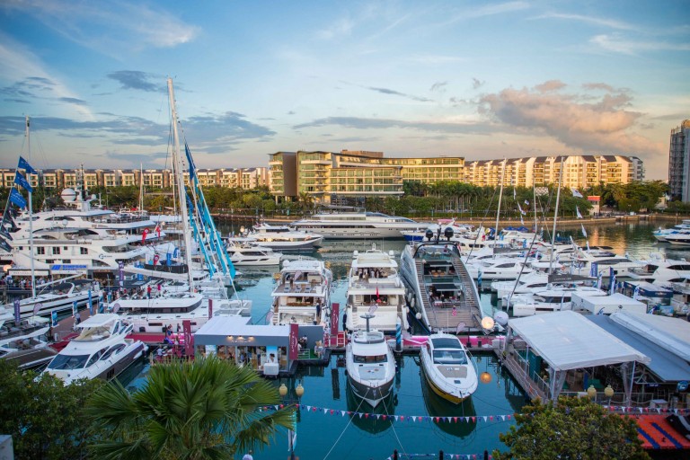 A marina filled with numerous yachts and boats moored in the water, with modern buildings and flats in the background under a blue sky with some clouds. Palm trees and colourful flags line the waterfront.