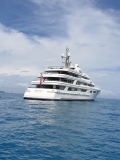 A large white luxury yacht is anchored on calm blue water under a partly cloudy sky. The yacht has multiple decks and a red flag on its aft.