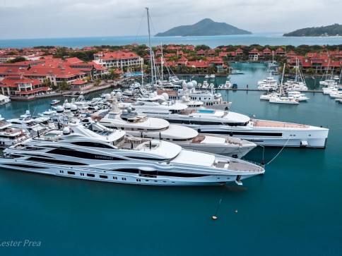 Aerial view of luxury yachts moored in a marina with red-roofed buildings, turquoise water, and a mountainous island in the background under a cloudy sky.