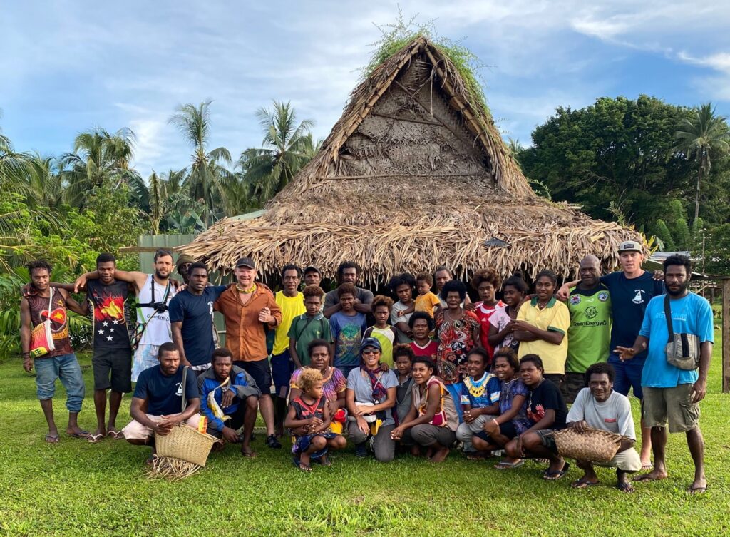 A group of people of various ages pose together in front of a traditional thatched-roof hut, surrounded by lush greenery and palm trees, on a bright day.