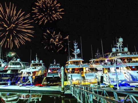 Several yachts are moored at a marina at night, brightly lit, with colourful fireworks bursting in the sky above, reflecting on the water.