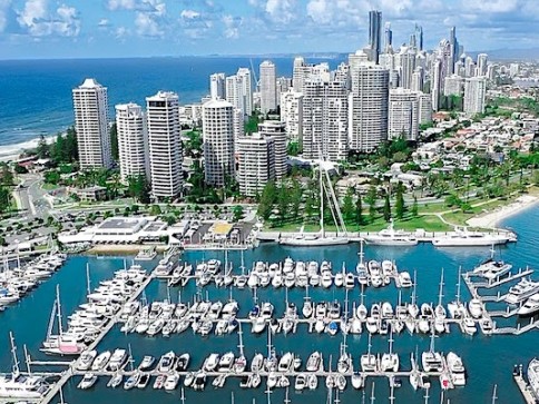 Aerial view of a marina filled with numerous yachts and boats, with a backdrop of tall modern skyscrapers and coastal buildings under a bright blue sky near the sea.