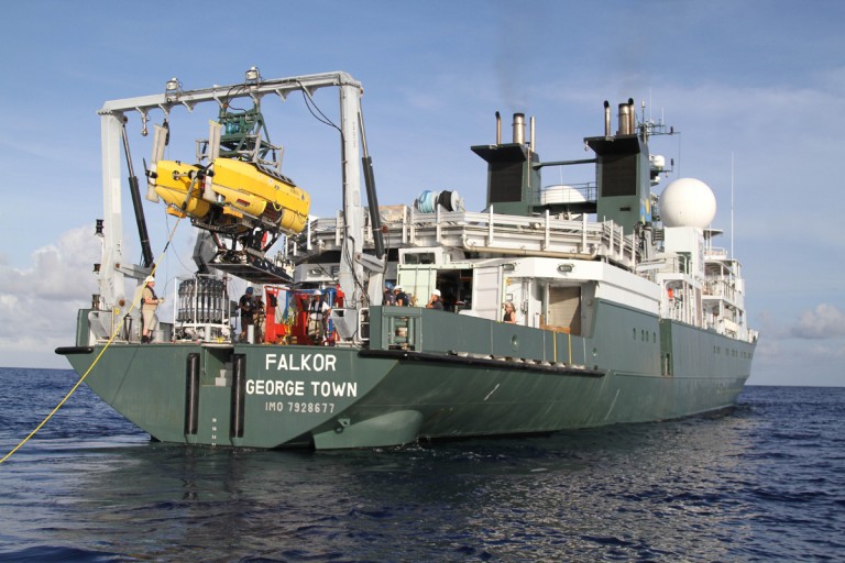 Research vessel Falkor floats on the ocean, with crew members operating equipment at the stern and a yellow underwater submersible being lowered into the water by a crane.