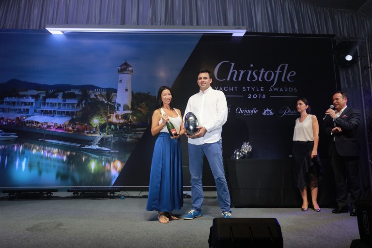 Two people stand centre stage holding silver awards at the Christofle Yacht Style Awards 2018, with a backdrop of a marina and lighthouse. Three others stand nearby, one speaking into a microphone.