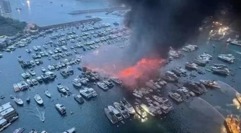 A large fire burns among boats moored in a crowded marina, sending thick black smoke into the sky as emergency crews spray water to control the blaze.