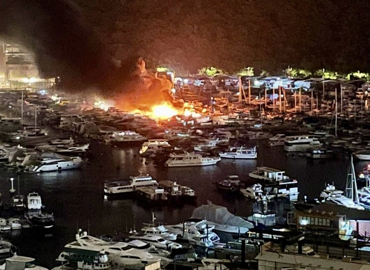 A nighttime view of a marina filled with boats, with flames and thick black smoke rising from a fire among the vessels, illuminating the surrounding area. Dense trees or hills are visible in the background.