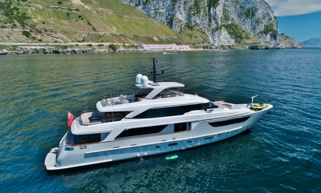 A large, modern white yacht floats on calm blue water near a rocky coastline with green hills and cliffs in the background. The yacht has multiple decks and outdoor seating areas.