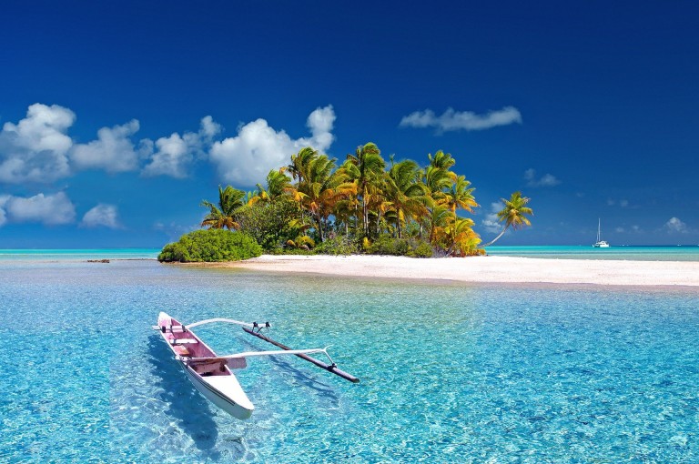 A small tropical island with palm trees sits in turquoise water under a clear blue sky. In the foreground, a white outrigger canoe floats on the calm sea, and a sailing boat is visible in the distance.