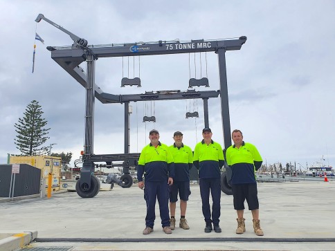 Four workers in high-visibility shirts stand in front of a large 75-tonne mobile boat hoist at a marina, with overcast skies and boats visible in the background.